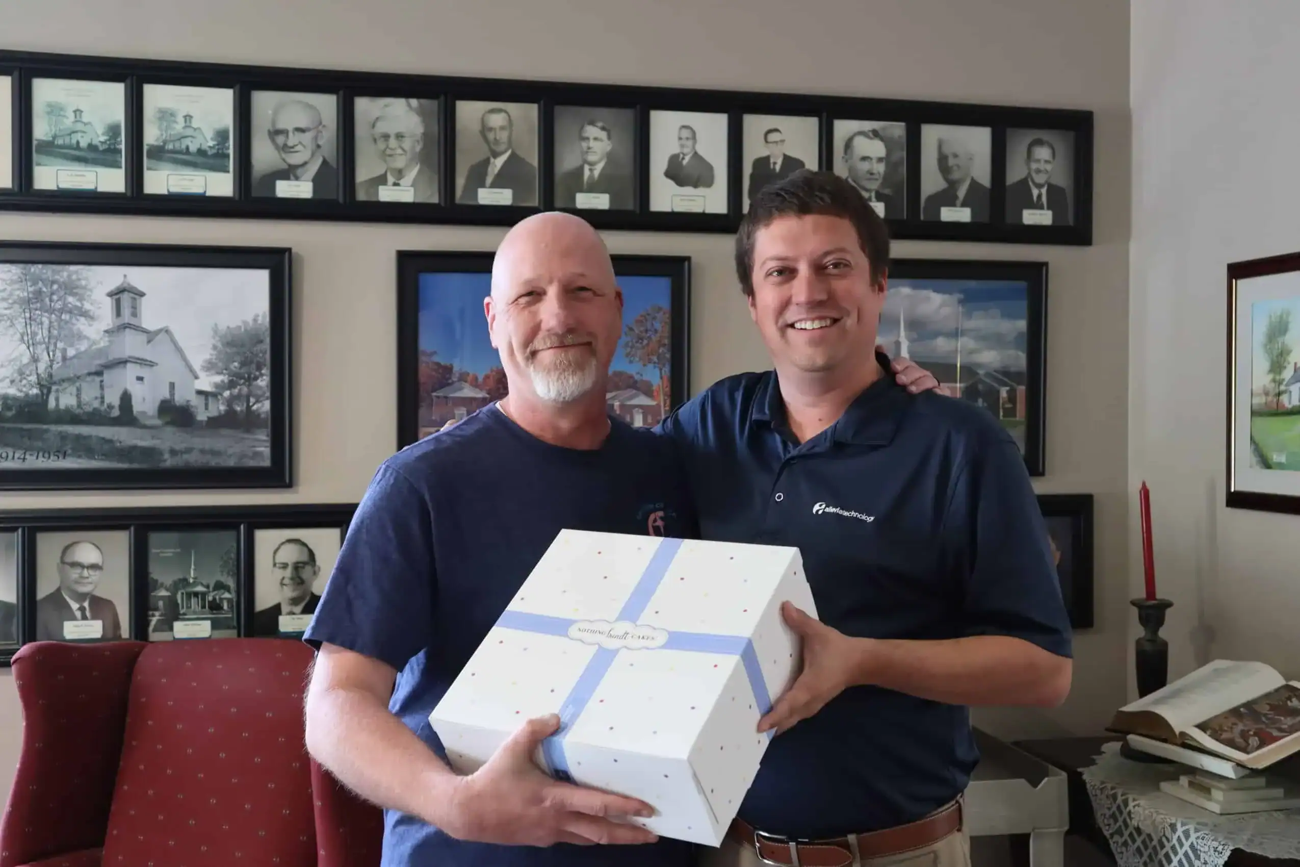 Two men smiling, holding a white gift box in an office setting.