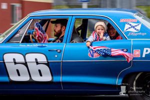 American race car celebrating patriotic pride with family in a vintage blue racing car, featuring American flags and racing decals.