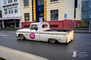 Rustic vintage pickup truck with "Good Home" sticker, part of Americarna event, showing classic American cars on New Zealand streets.