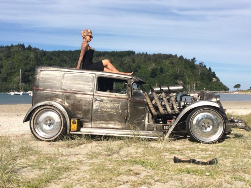 Vintage hot rod car on beach with woman sitting on roof, American car enthusiasts at Americarna event, classic custom vehicle photography, New Zealand coastline.