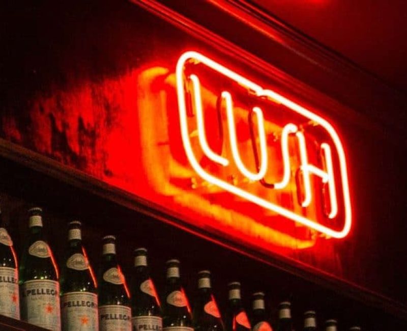 Neon "Open" sign lights up a vintage car show venue, with a row of beer bottles on display nearby.