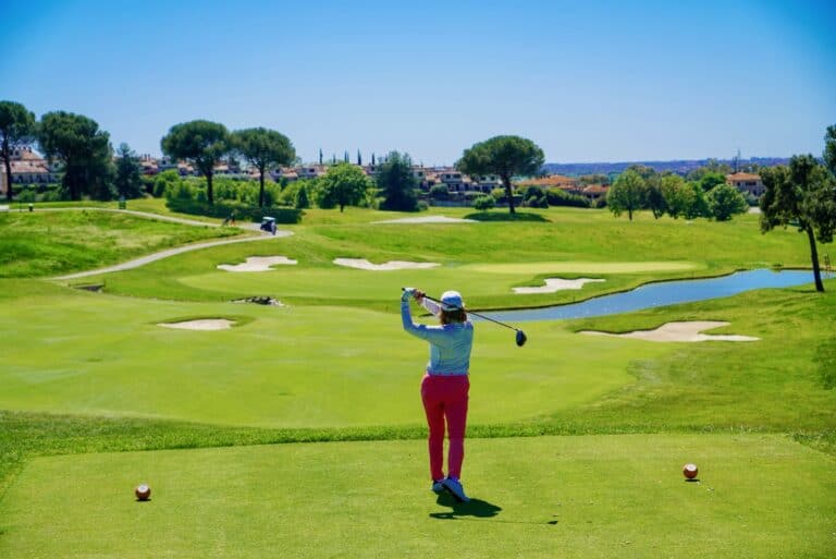 Woman playing golf on a sunny course with trees and water hazards.
