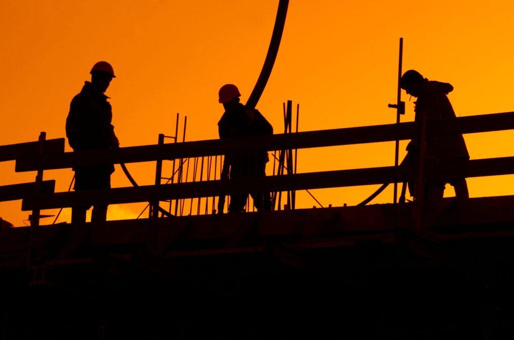 Travailleurs de la construction en silhouette contre un ciel orange au coucher du soleil, portant des casques de sécurité sur un chantier.