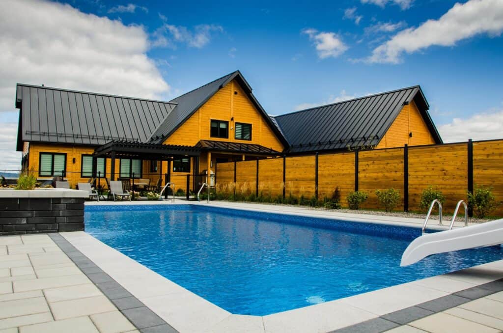 Piscine creusée avec escalier en métal, maison en bois avec toiture en métal noir, clôture en bois, aménagement extérieur, maison moderne, espace détente extérieur, conception de piscine résidentielle.