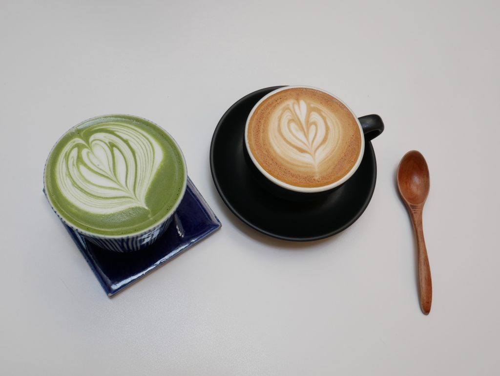 A lush green matcha latte with latte art on a blue saucer, alongside a classic cappuccino with latte.