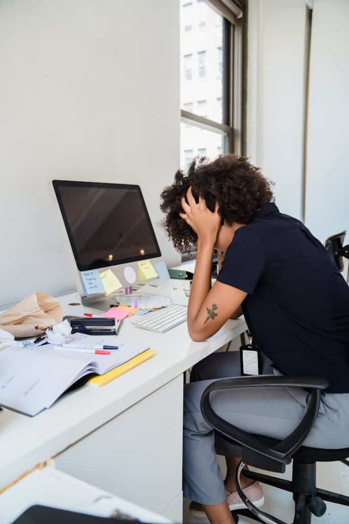 Frustrated Woman Sitting by Desk wearing a black shirt holding her heads in her hand. This can represent what it can look like for a frustrated neurodiverse employee