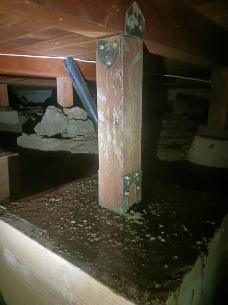 Wooden post supporting a floor beam in a dark crawl space.