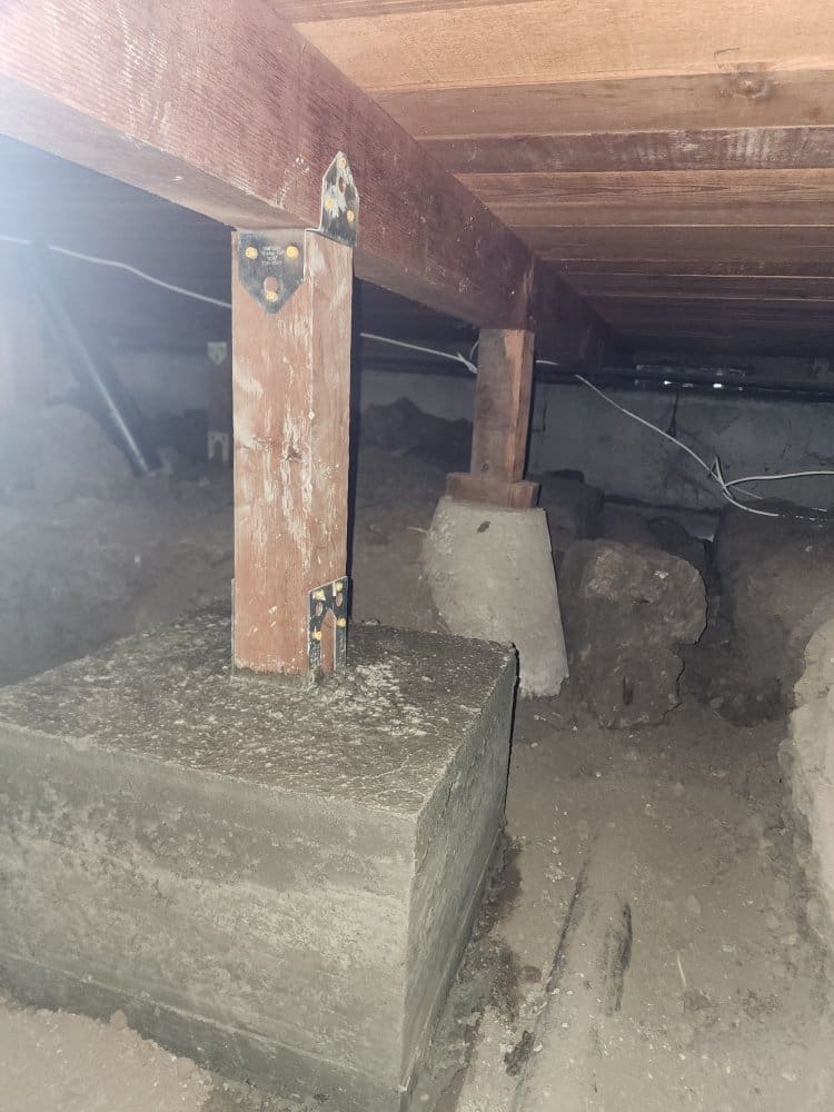 Crawl space with wooden beams, concrete piers, and dirt floor.