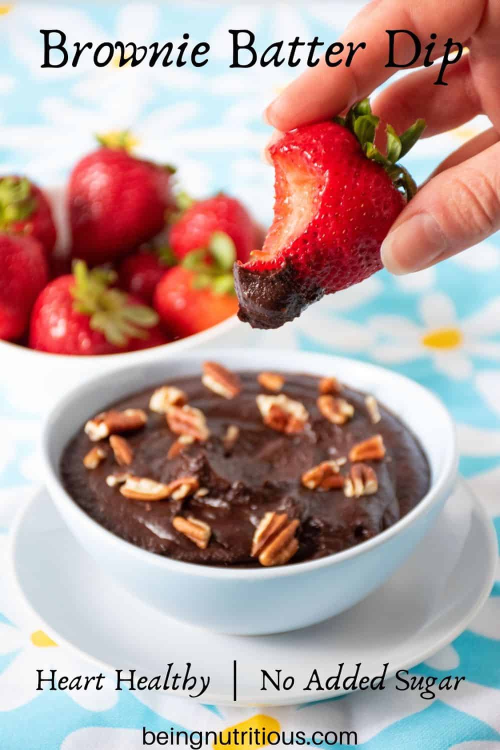 Brownie batter dip in a bowl with a bowl of strawberries behind. A hand holding a strawberry with dip on it and a bit taken out in the foreground.