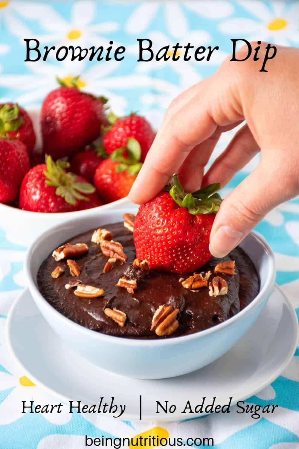 Brownie batter dip in a bowl with pecan pieces on top, and a hand dipping a strawberry in.