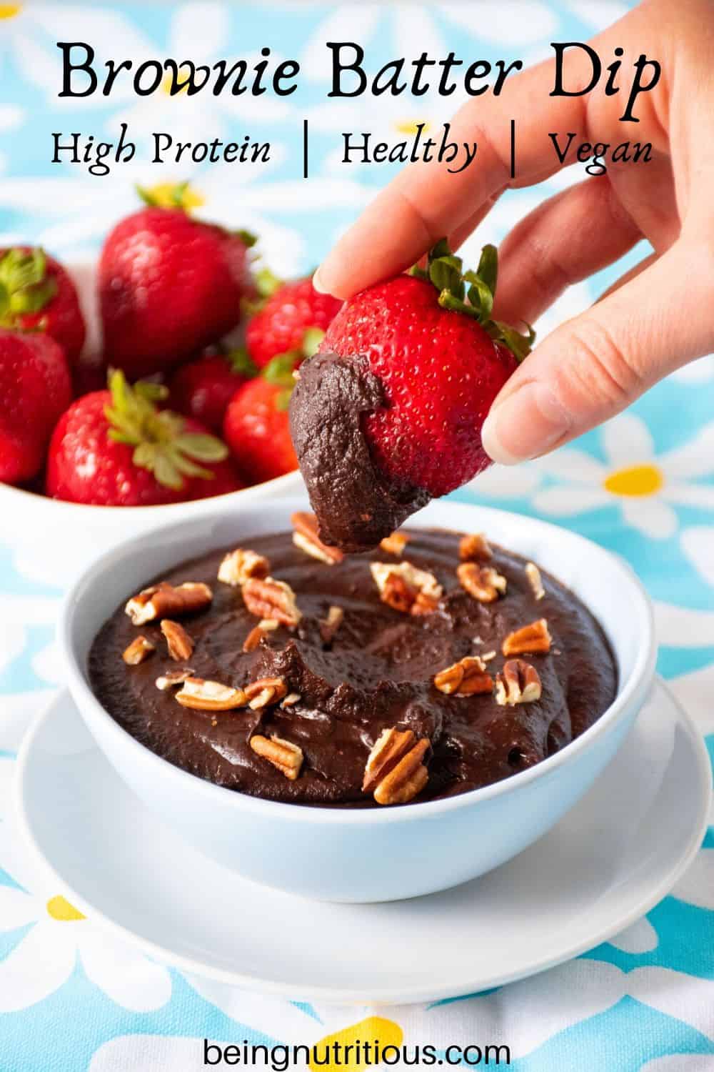 Brownie batter dip in a bowl, with a hand holding a strawberry with the dip on it.