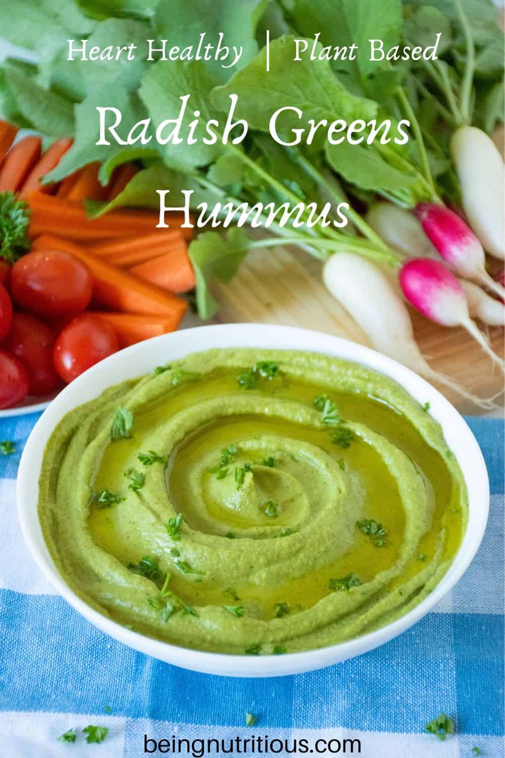 Radish greens hummus in a white bowl with cherry tomatoes, carrot sticks and radishes in the background.