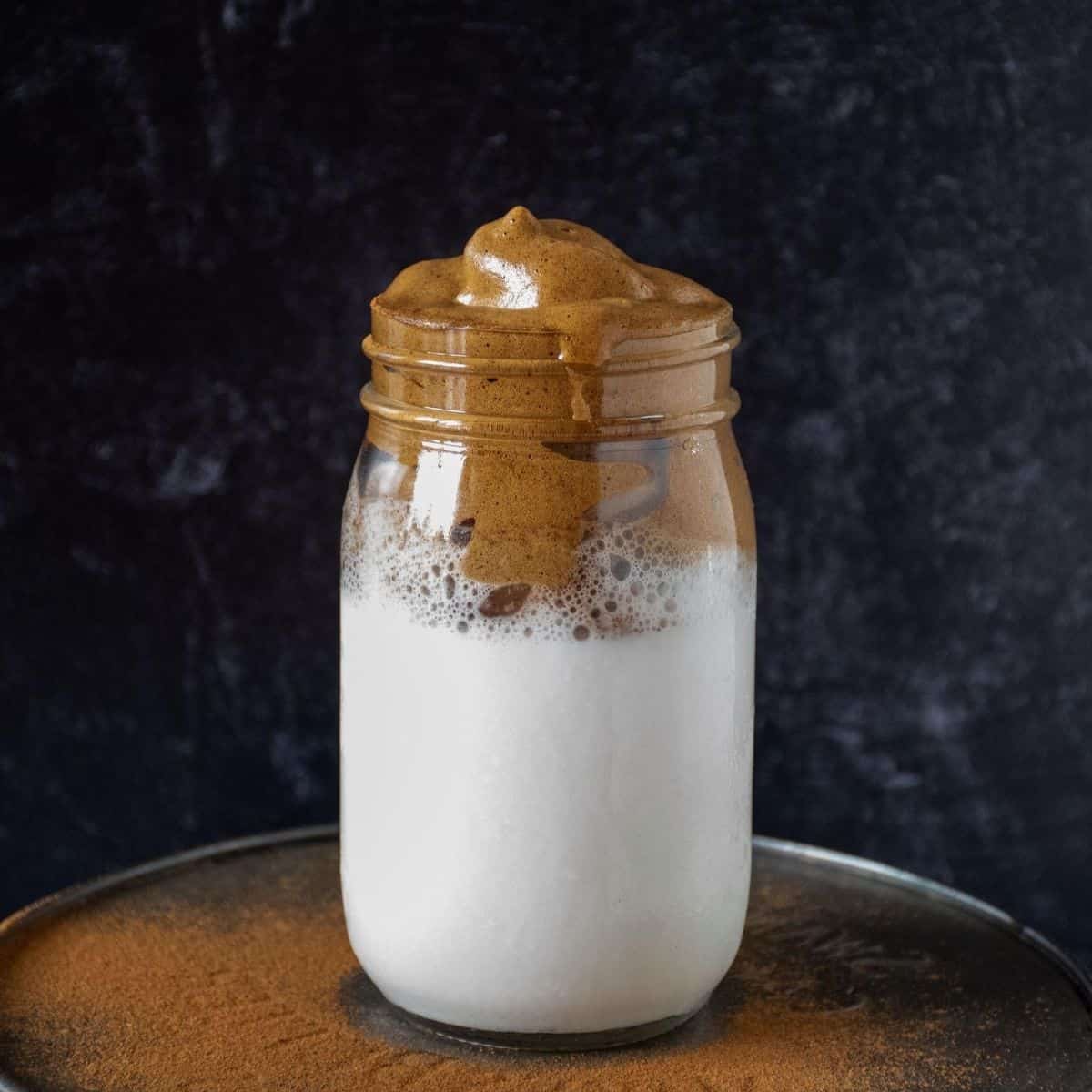 Frothy, whipped coffee, over iced almond milk, in a Mason jar in front of a dark background.