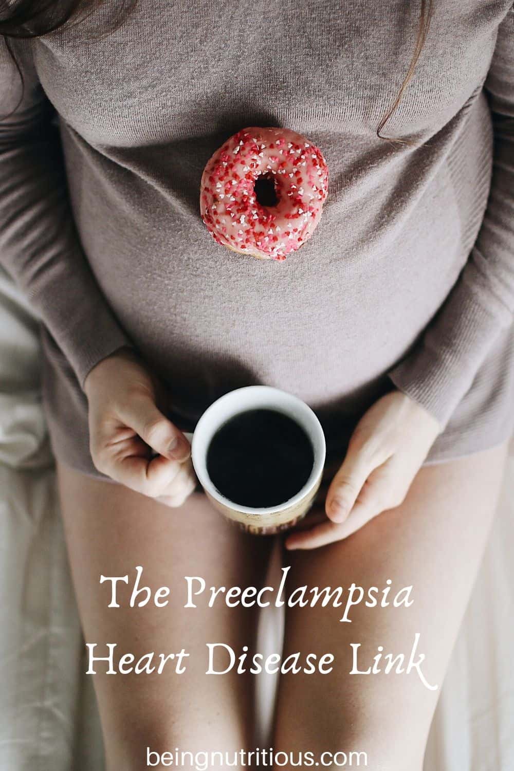 overhead shot of a pregnant woman from the chest down, wearing a gray sweater with a doughnut on her belly, holding a cup of coffee.