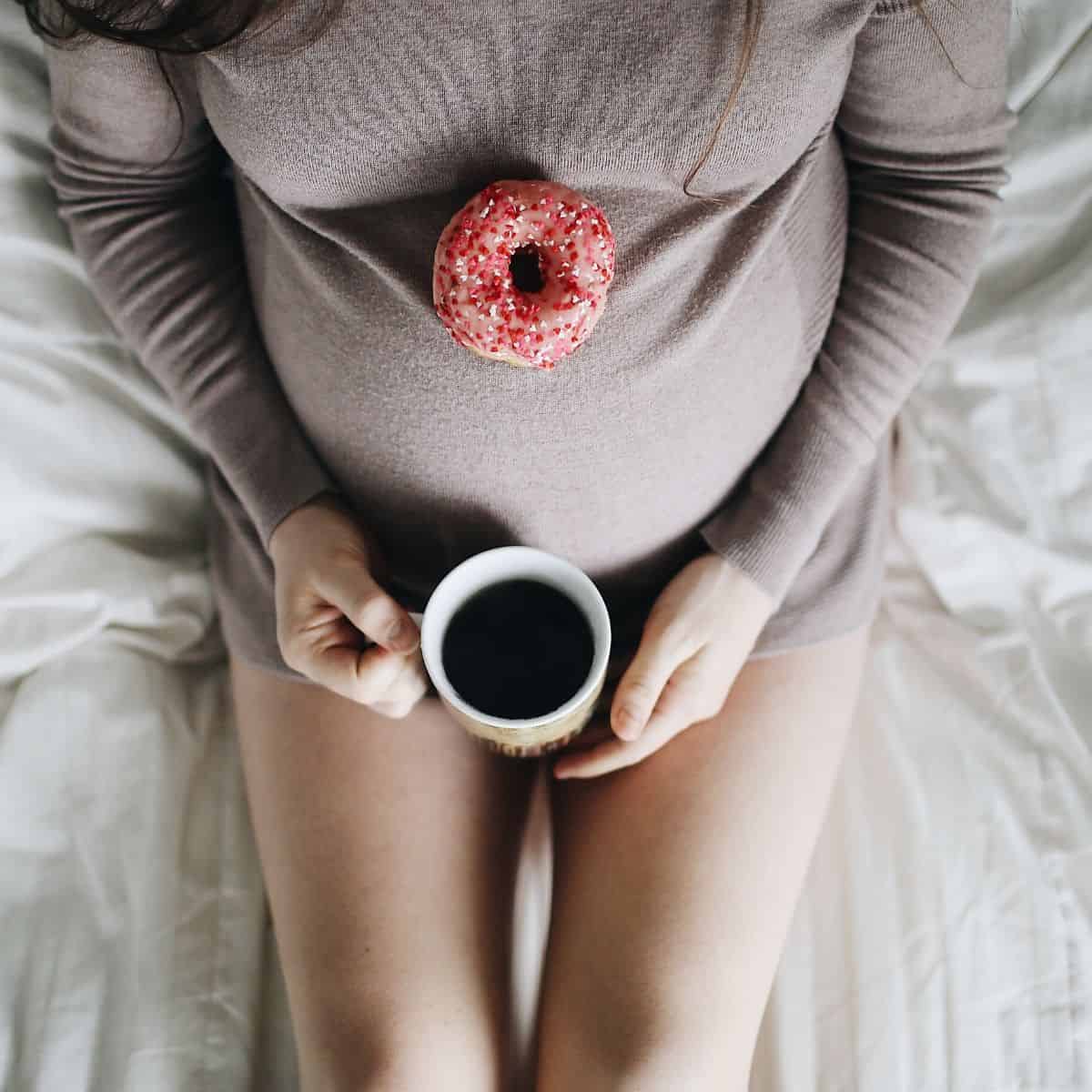 pregnant woman from the chest down, wearing a gray sweater, holding a cup of coffee with a doughnut sitting on her belly