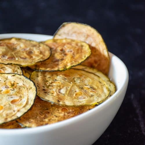 Zucchini chips in a white bowl in front of dark background.