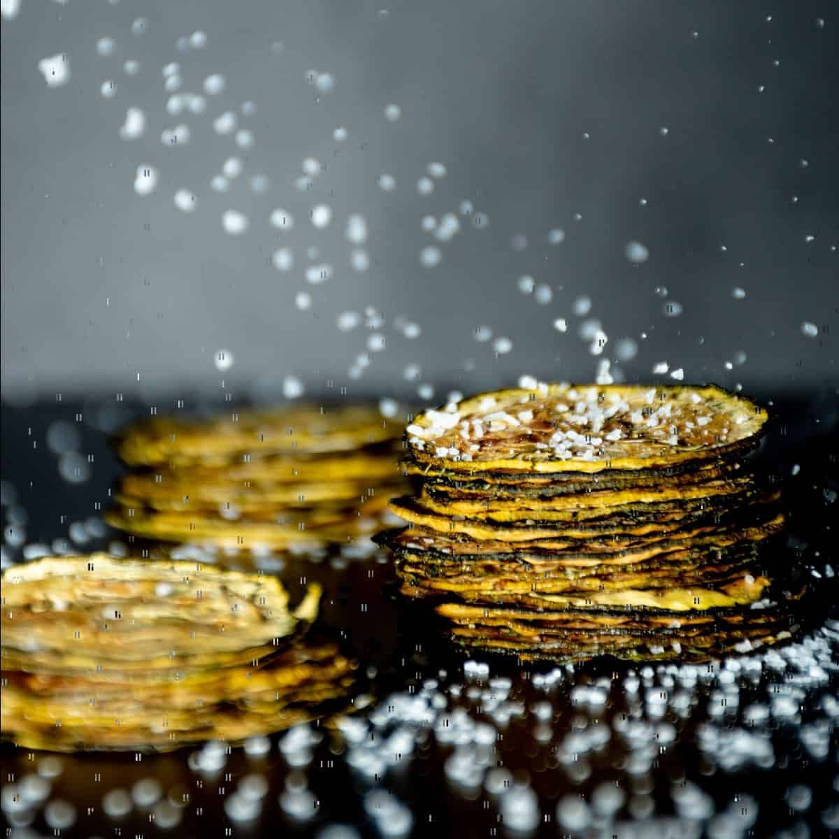 Square image of 3 stacks of zucchini chips, with salt crystals falling all around.