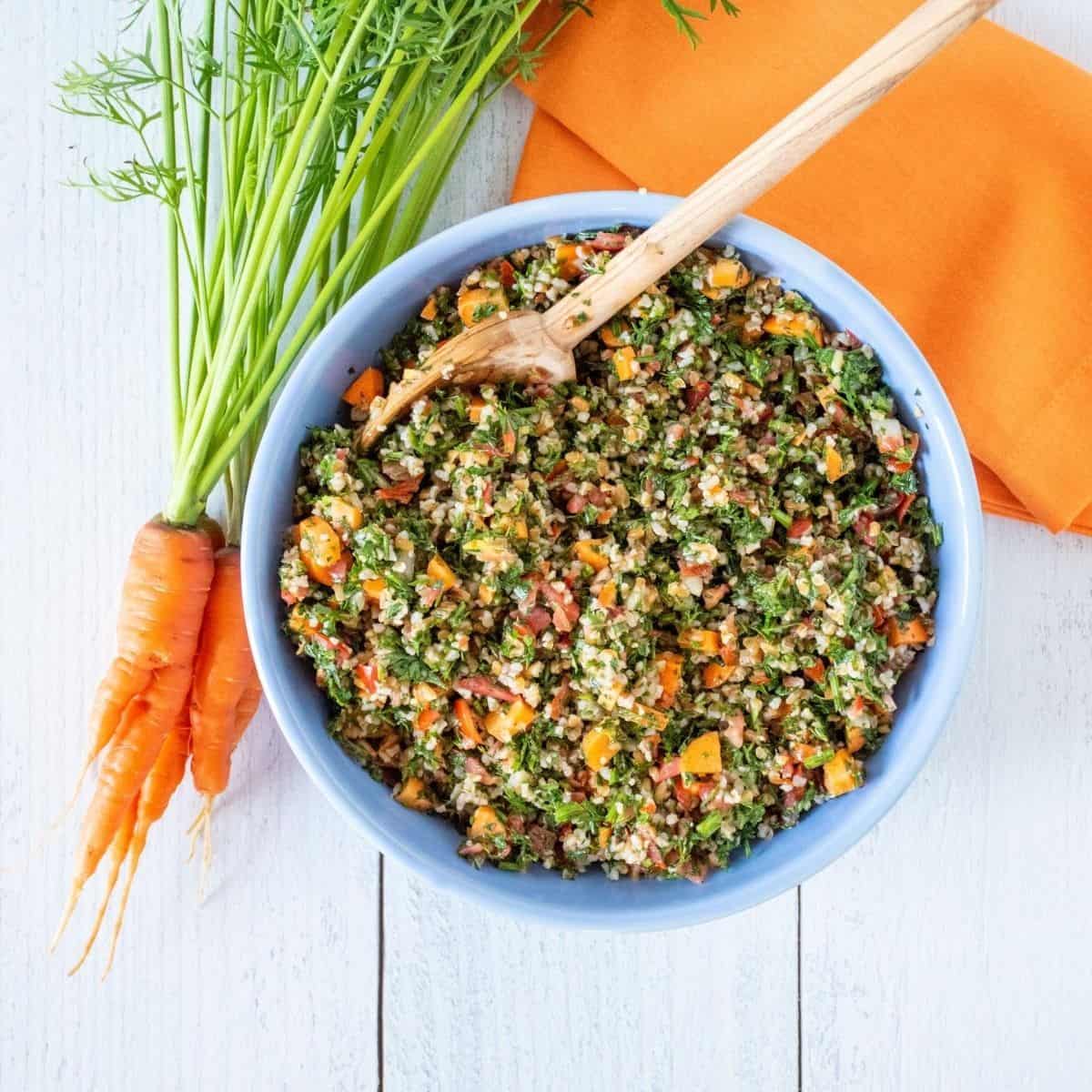 Overhead shot of carrot top tabbouleh in a blue bowl, half of the bowl shown, with a bunch of carrots with the tops still attached lying beside the bowl.