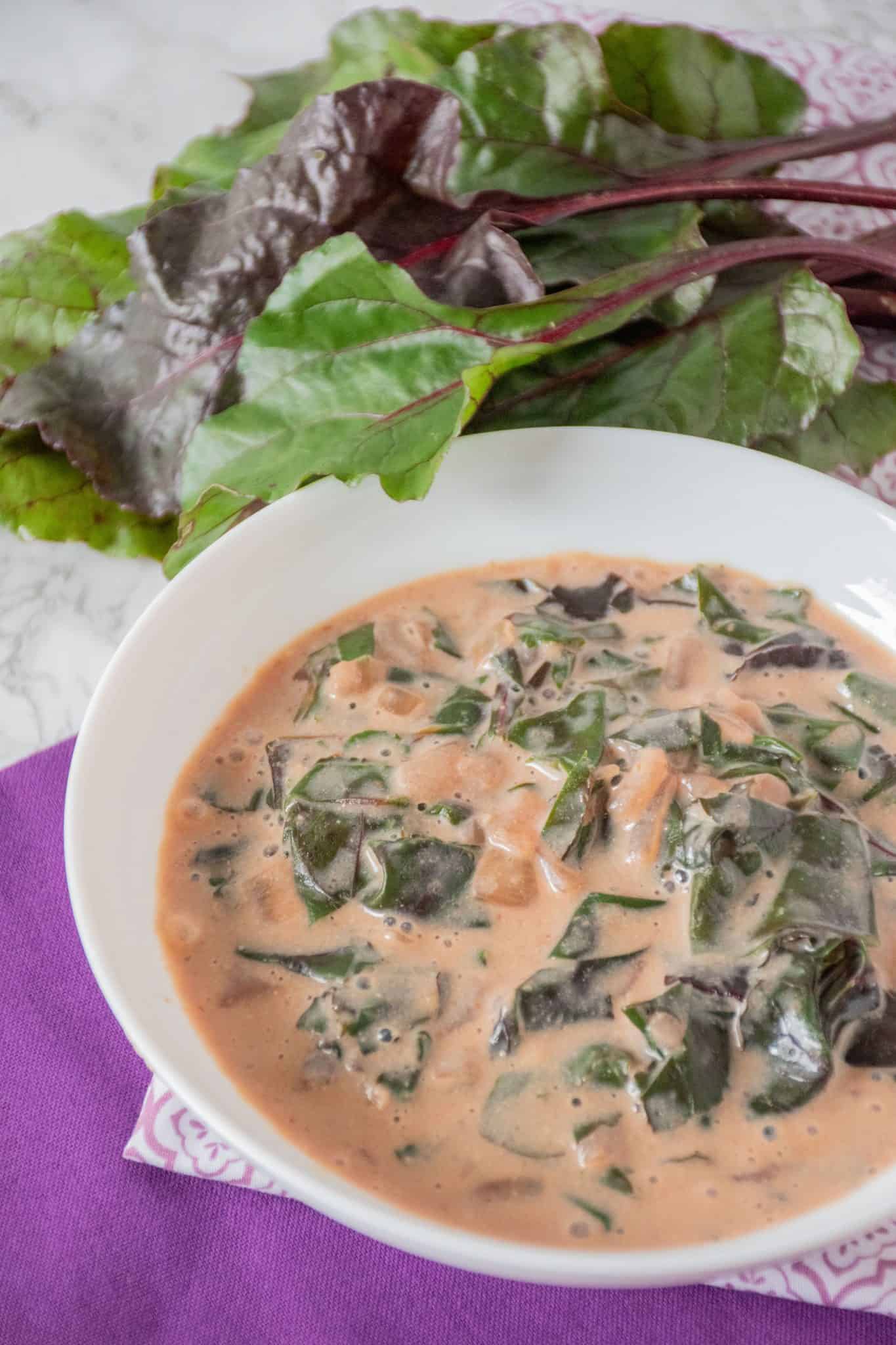 Bowl of braised greens in a light pink broth. A bunch of beet greens visible behind the bowl.