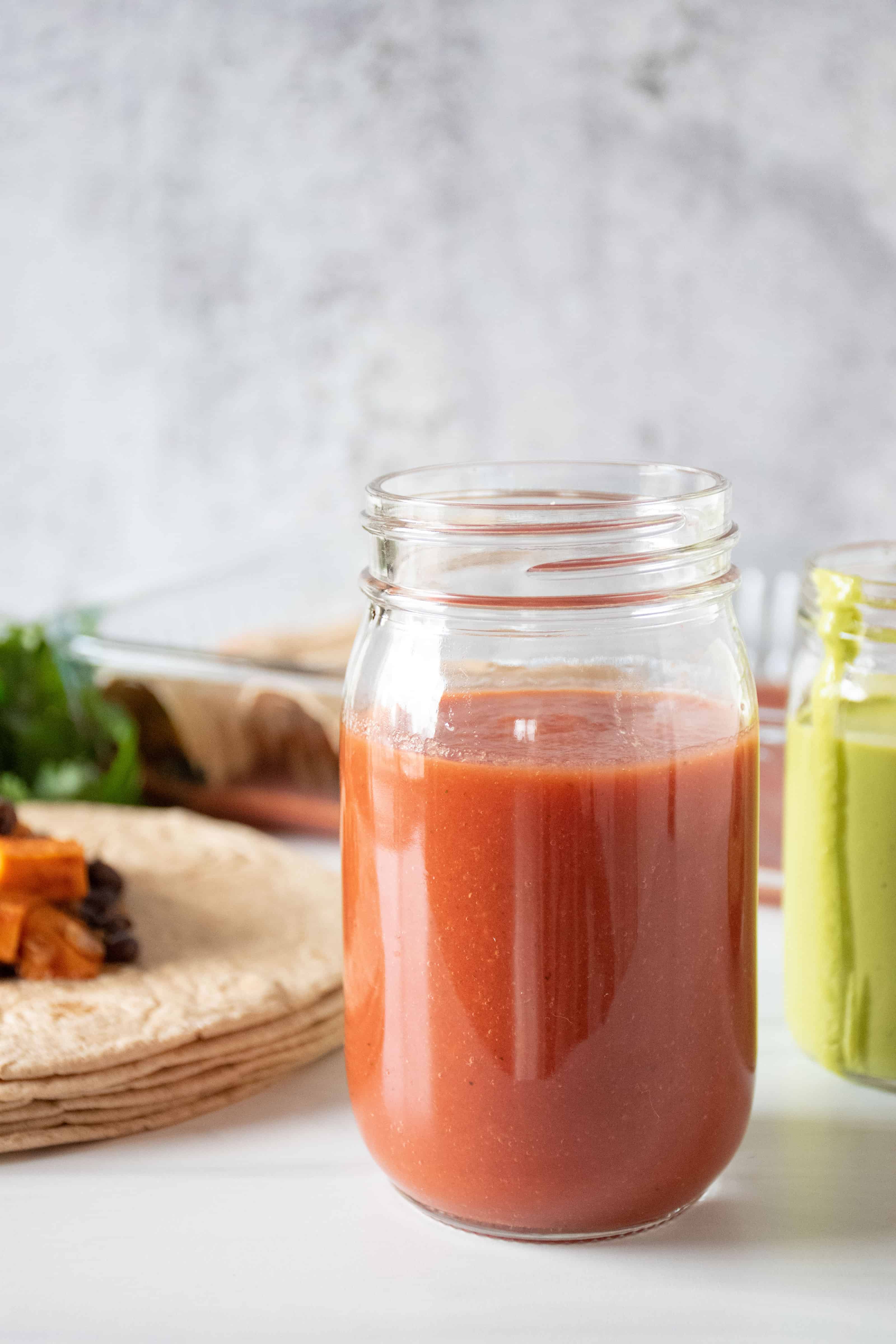 Red sauce in a jar, with enchiladas being made in the background. Text overlay: Fast and Easy! Low Sodium Enchilada Sauce