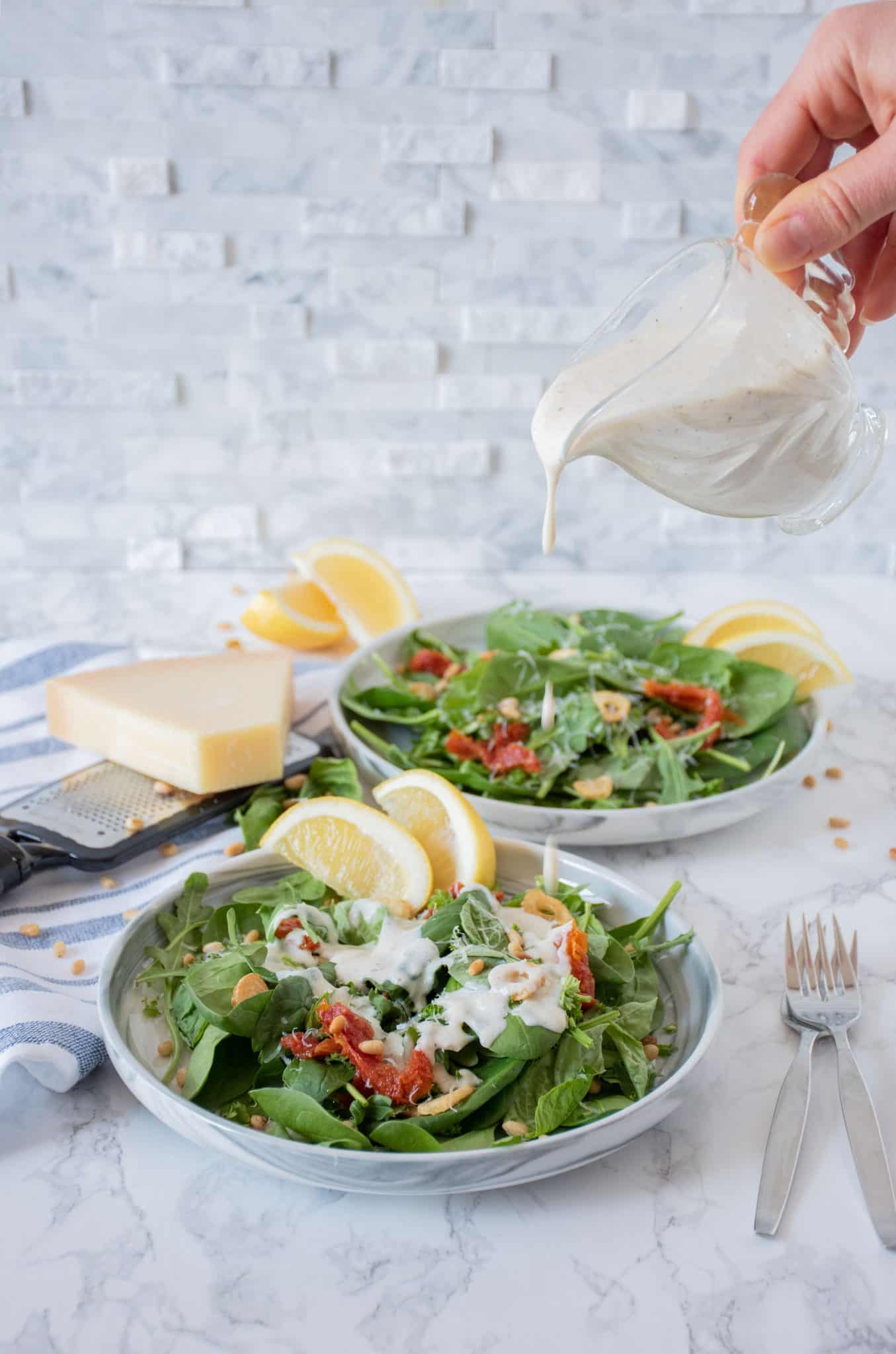 Plate of salad with sundried tomatoes, pine nuts, fried garlic, and dressing. being drizzled over
