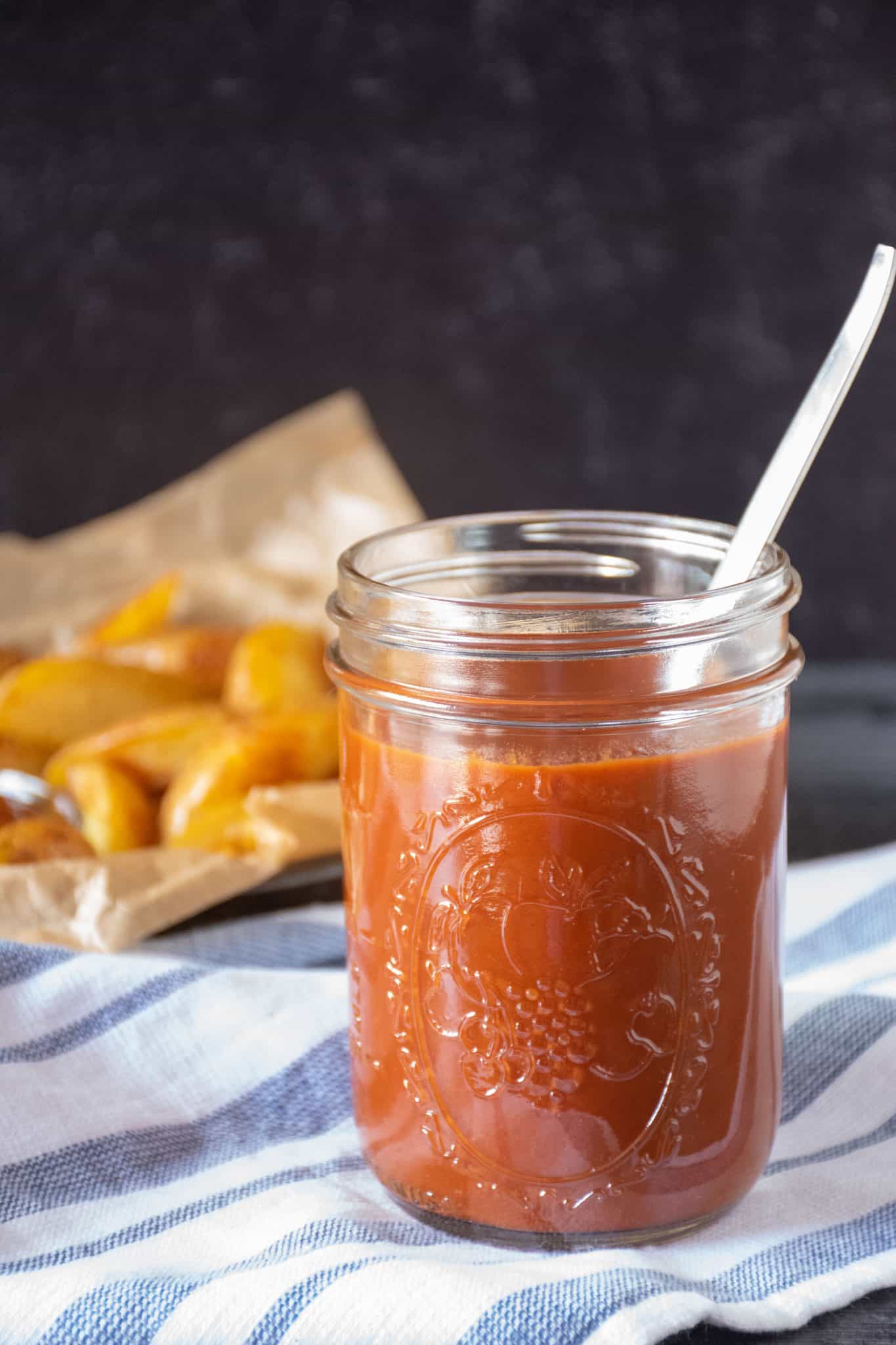 Jar of ketchup, with basket of potato wedges behind.