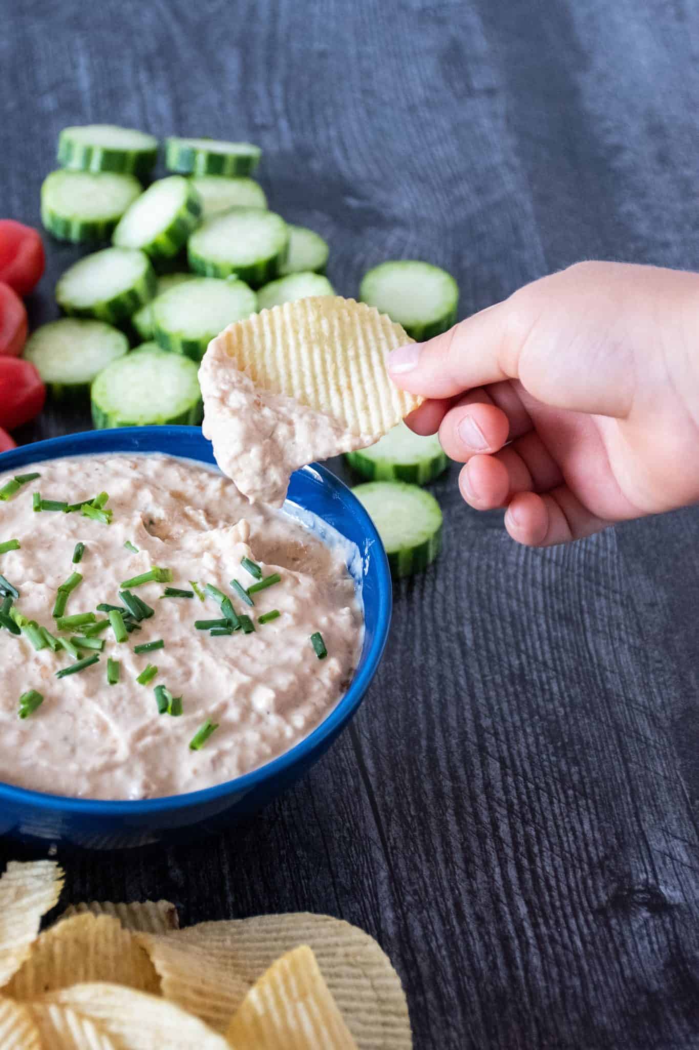 Bowl of dip, surrounded by chips, cucumber slices, and baby tomatoes. Hand holding a chip with dip on it above bowl.