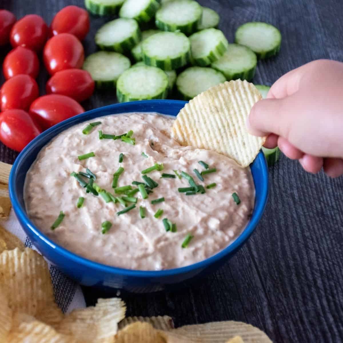Bowl of dip, surrounded by chips, cucumber slices, and baby tomatoes. Hand holding a chip with dipping it.