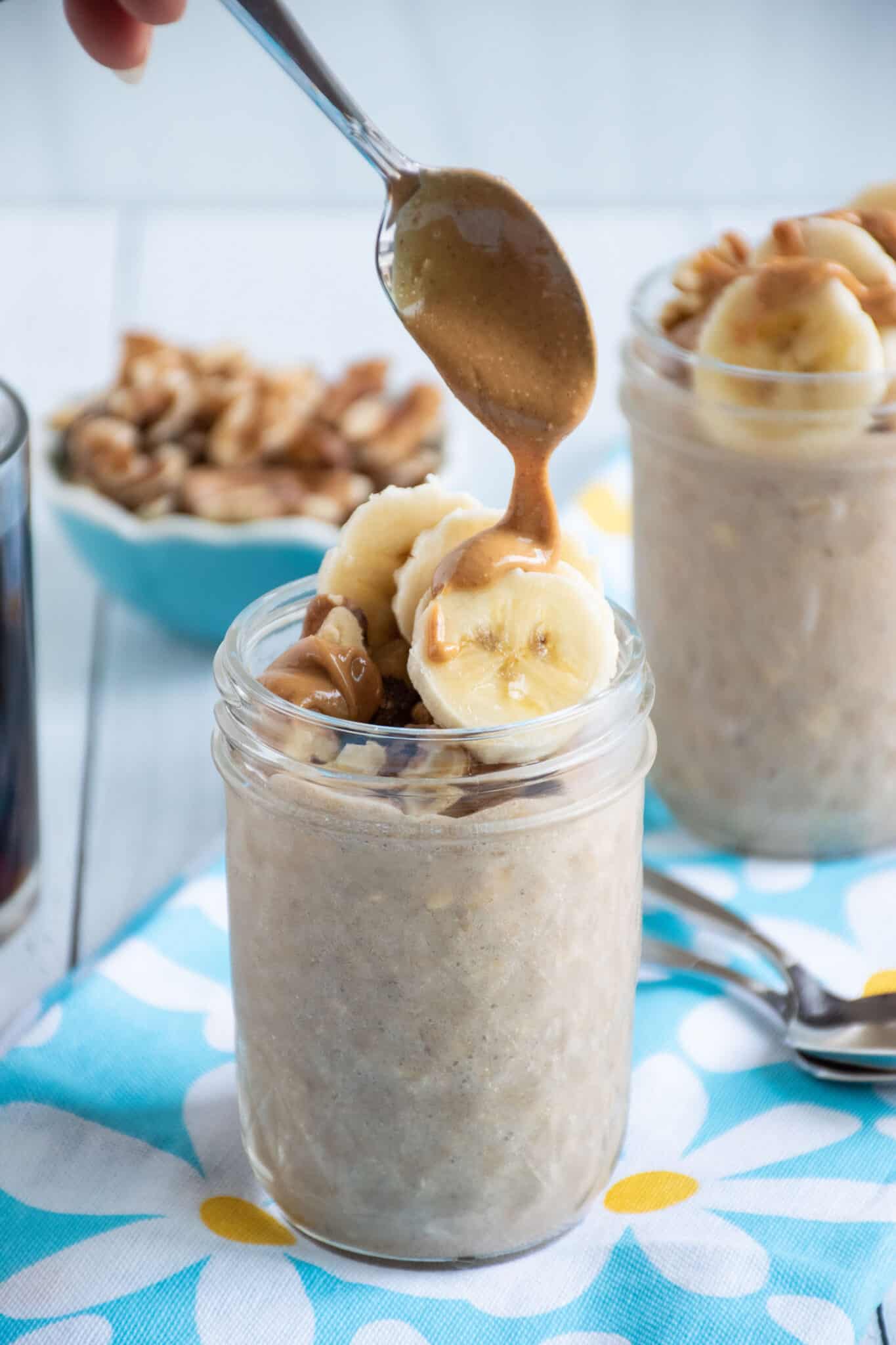 Small mason jar of oatmeal with walnuts and banana slices on top, being drizzled with peanut butter.
