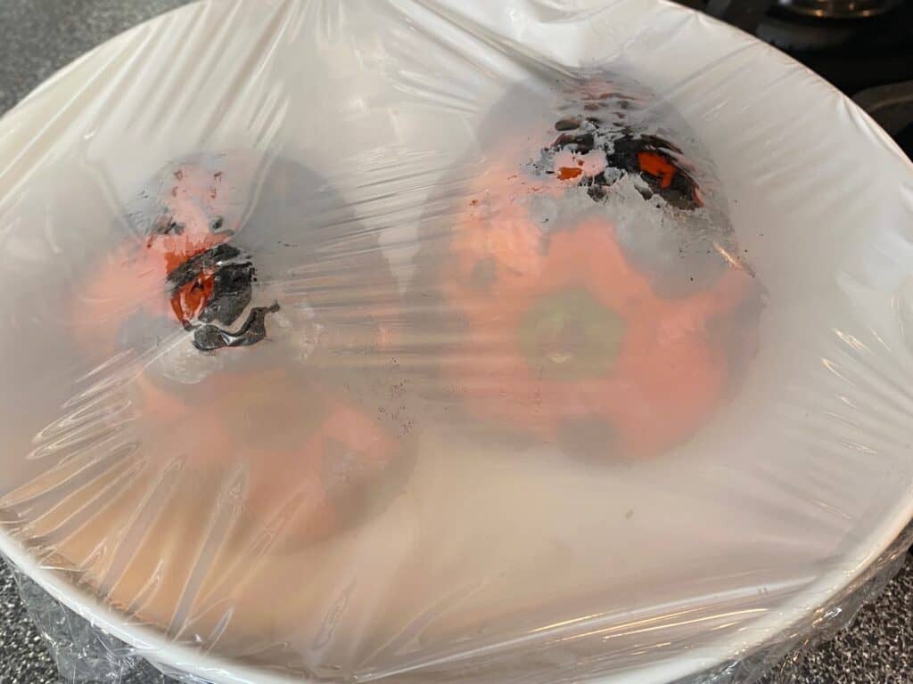 Charred red bell peppers in a bowl, covered with plastic wrap.