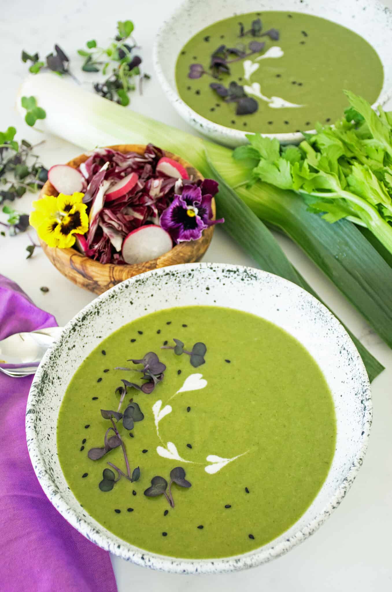 A bowl of celery soup, garnished with microgreens, black sesame seeds and yogurt. A side salad, another bowl of soup, and a fresh leek, celery, and microgreens are scattered about the background.