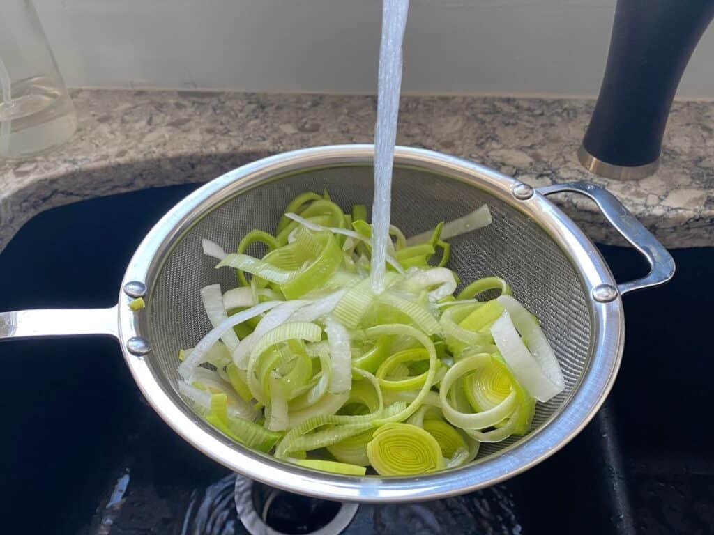 Sliced leeks in a mesh strainer, with water pouring over.