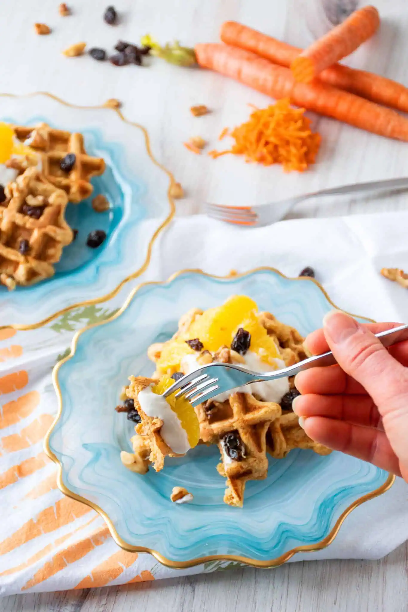 Plate of three small waffles, topped with yogurt, oranges, raisins, and walnuts. A hand is holding a fork with a waffle slice, some yogurt, and an orange slice on it in front of the plate.