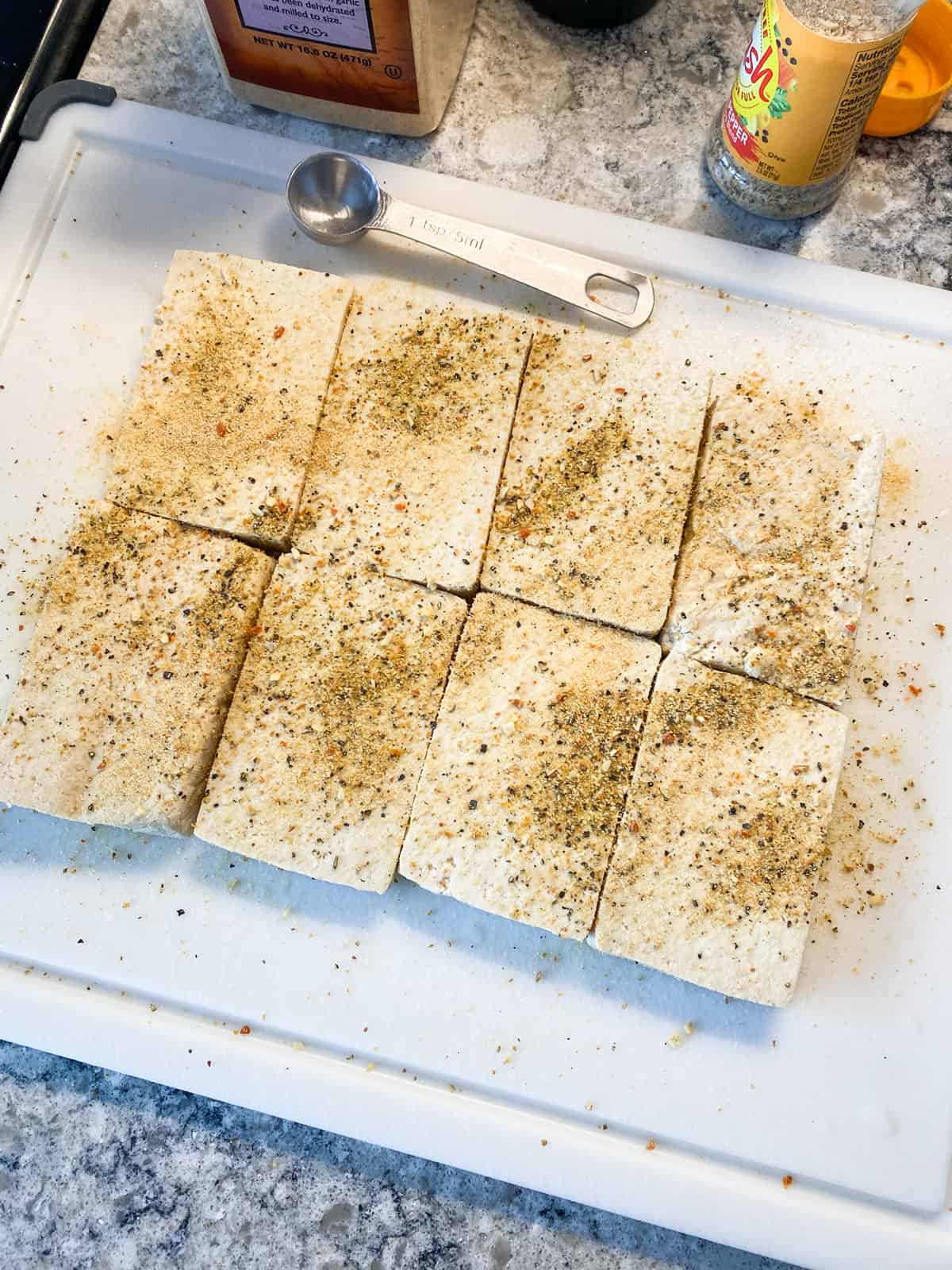 Slices of tofu on a cutting board, with seasonings sprinkled on them.