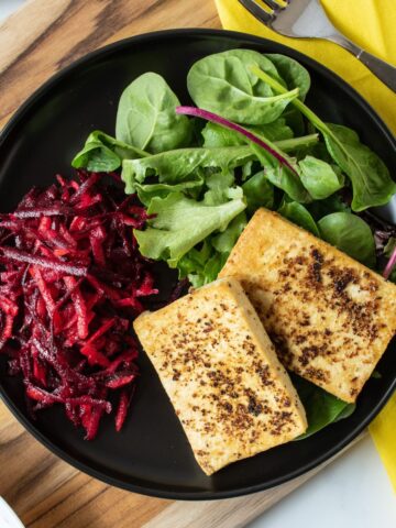 Overhead image of a plate with 2 slices of cooked tofu over a bed of greens, with a side of shredded beet salad.