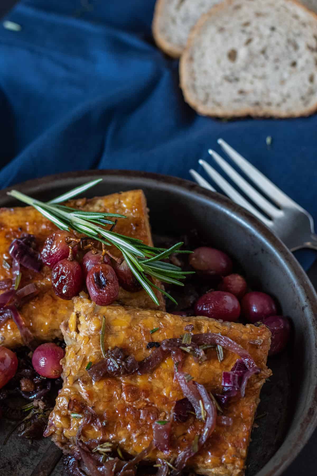 Metal dish with tempeh, caramelized onions, and grapes in it. Garnished with fresh rosemary.