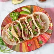 Five tacos lined up on a plate, surrounded by fixings.