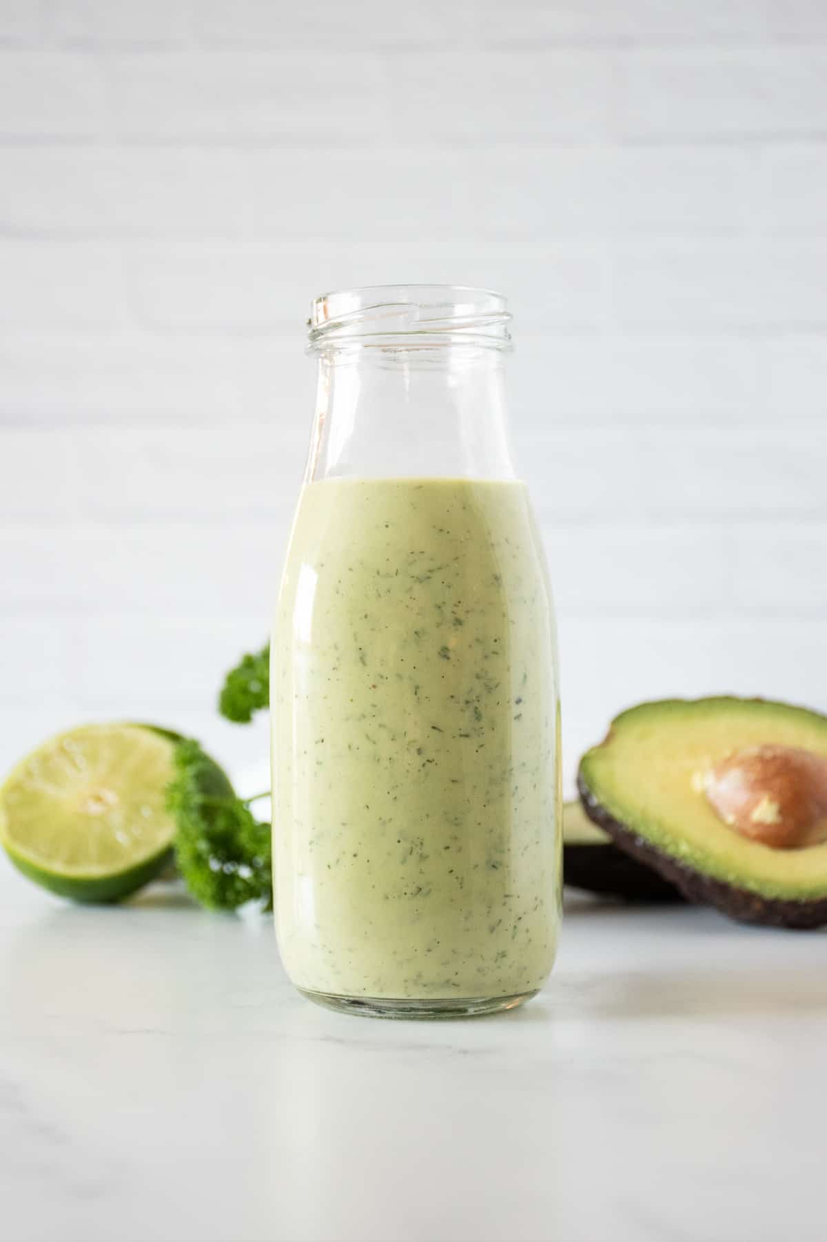 Jar of green colored salad dressing, with avocado, limes, and parsley in the background.