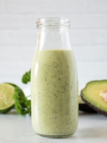 Jar of avocado salad dressing, with avocado, limes, and parsley in the background.
