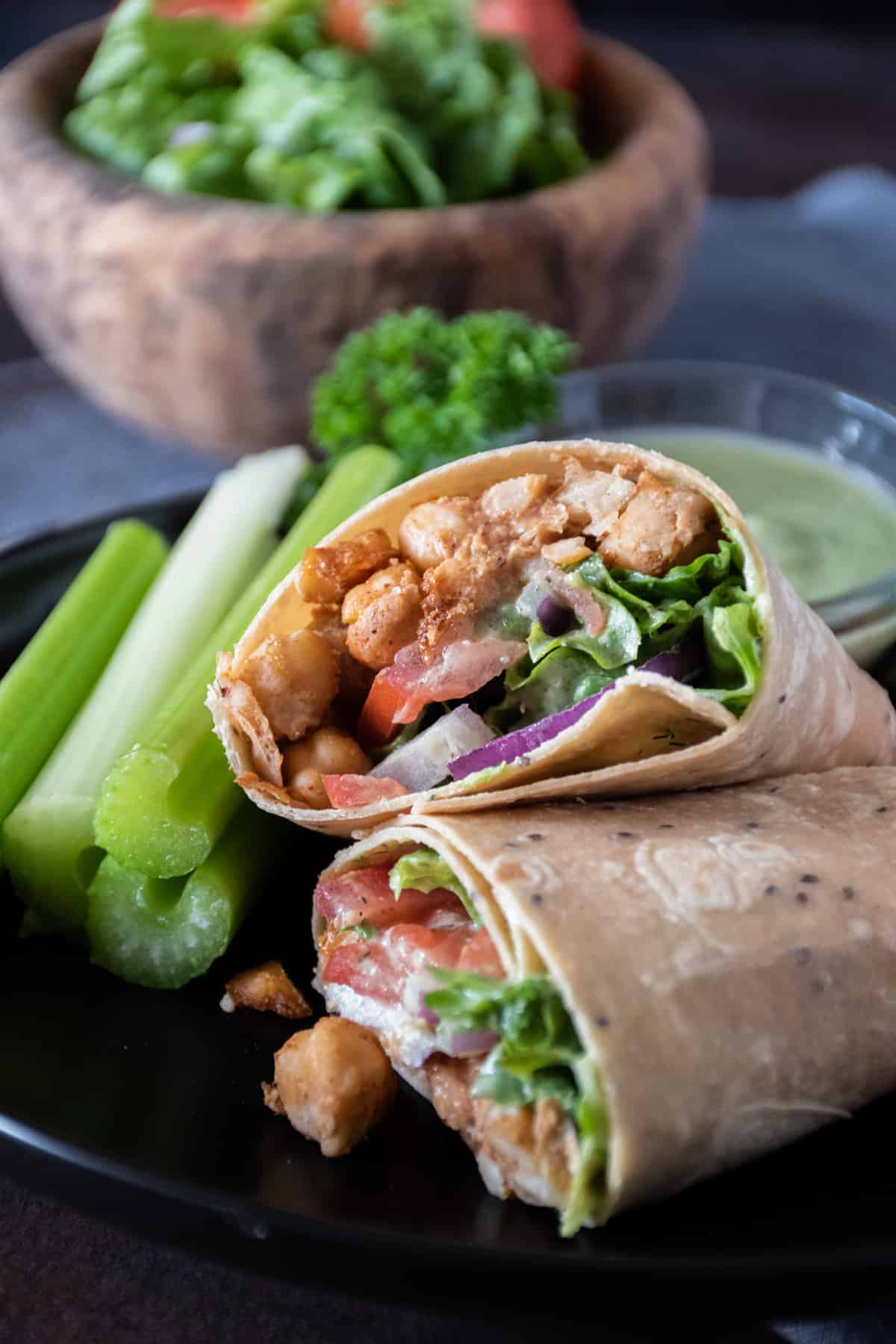 Wrap with chickpeas, lettuce, tomato and onion, cut in half, on a plate with celery sticks. A small bowl of salad in the background.