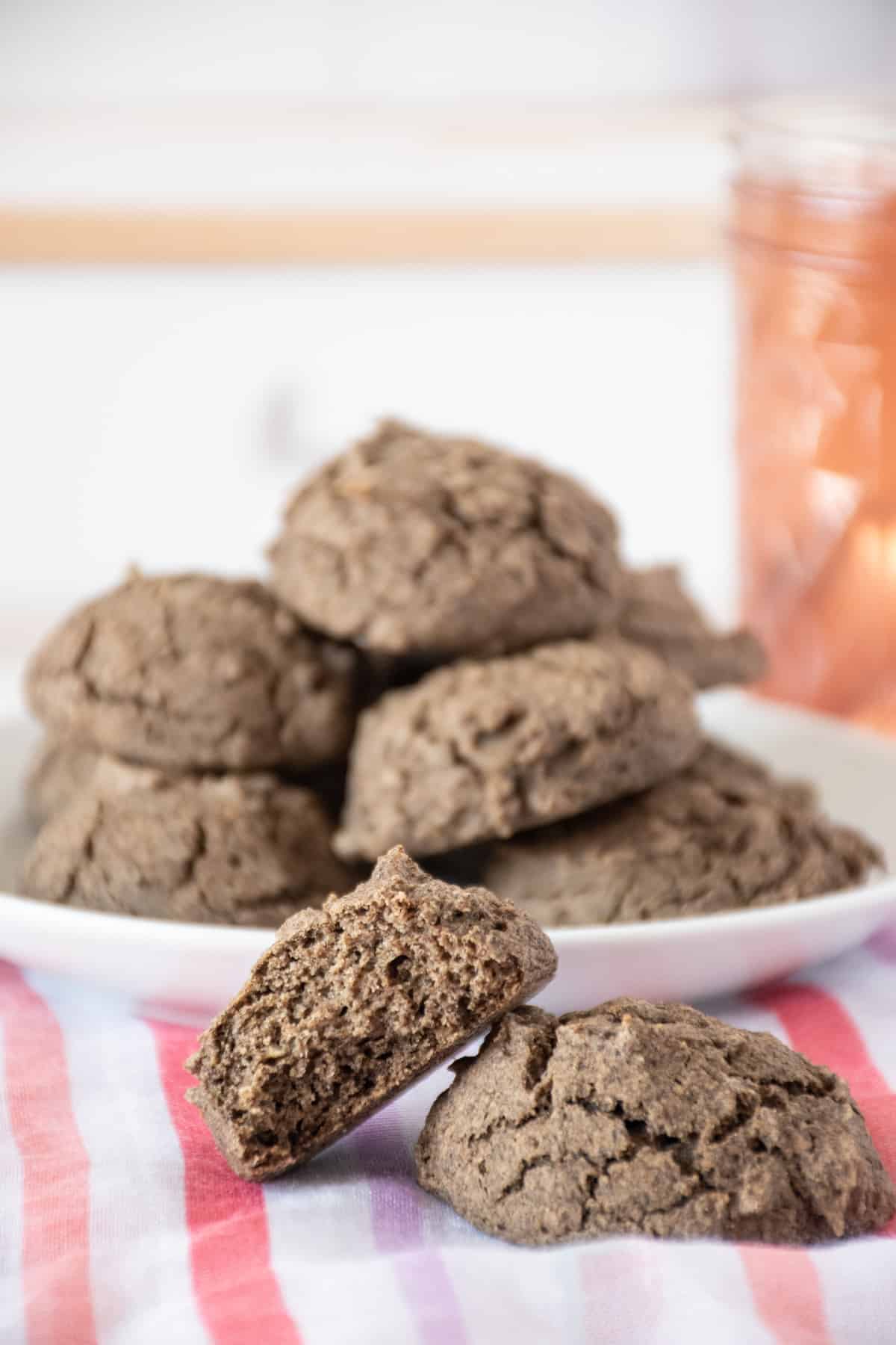 Two buckwheat biscuits, one with a bite taken out. A plate with more biscuits is in the background, and a small jar of jam.