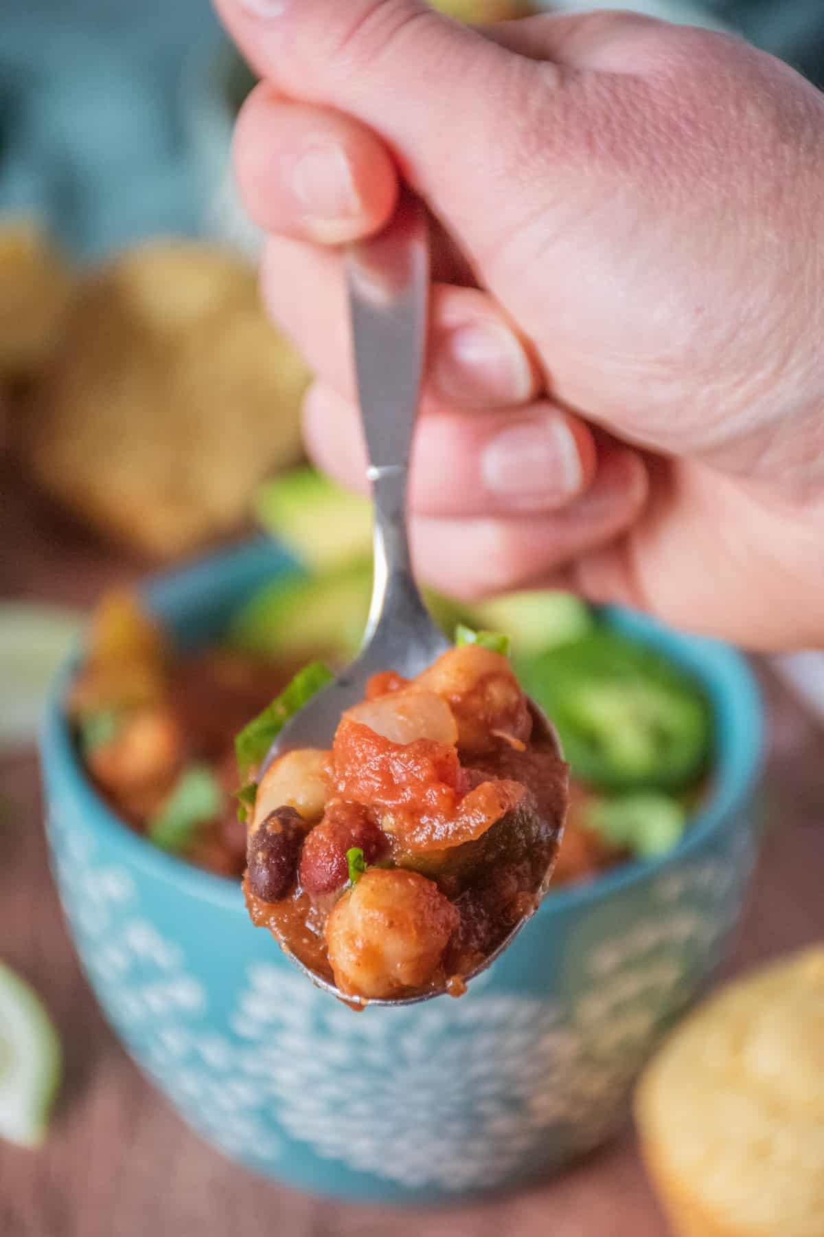 A hand holding a spoon with three bean chili on it. Bowl of chili visible in the background.