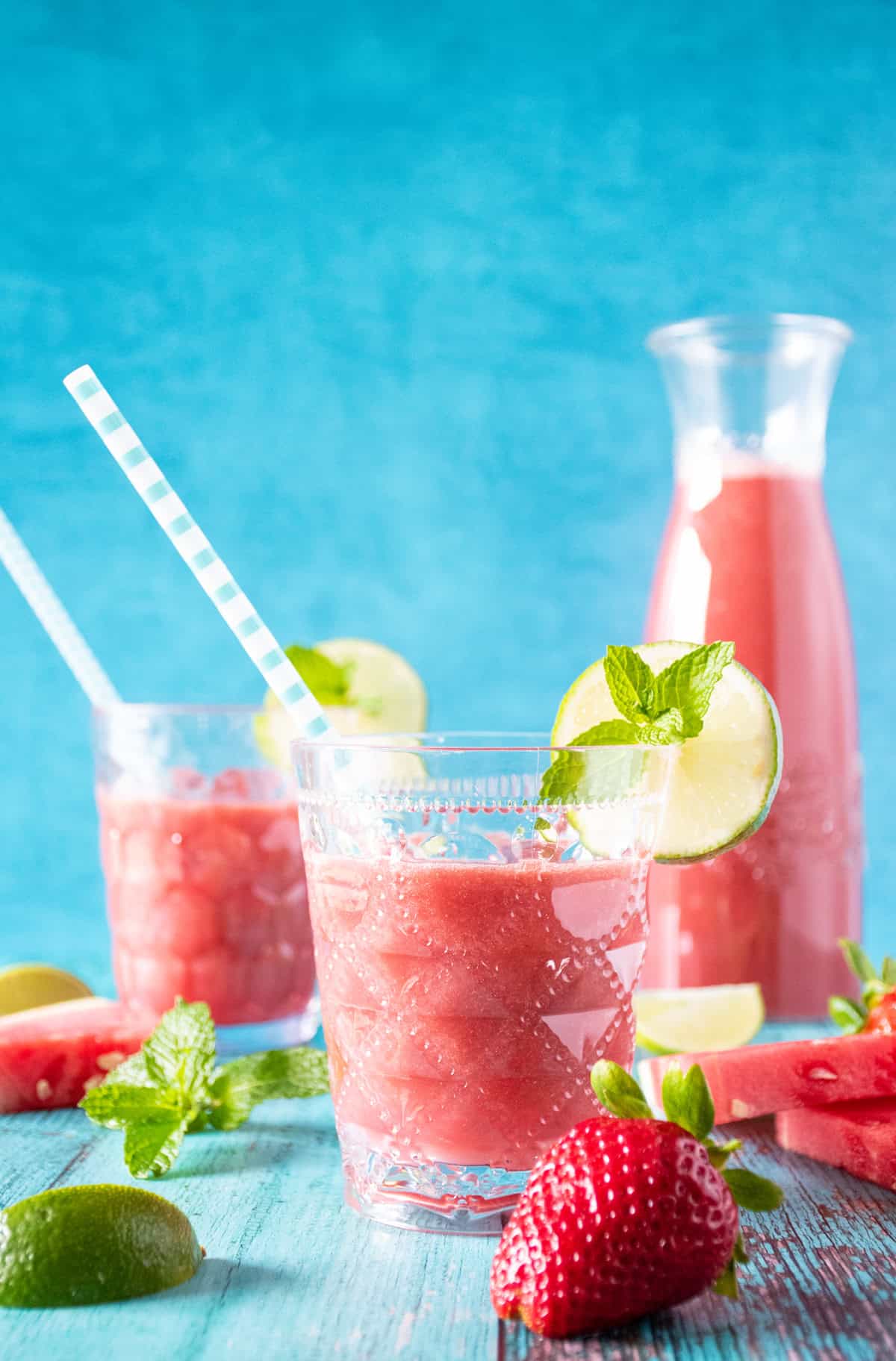 Glasses of agua fresca, with strawberries, watermelon slices, limes and mint scattered around. Carafe of beverage in background.