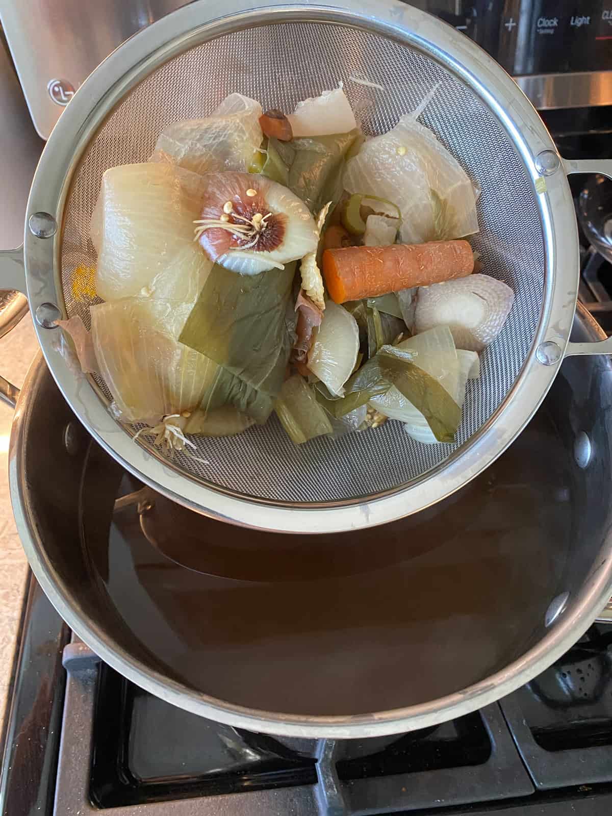 Vegetable stock, strained, in a stock pot. Vegetable scraps in a strainer.