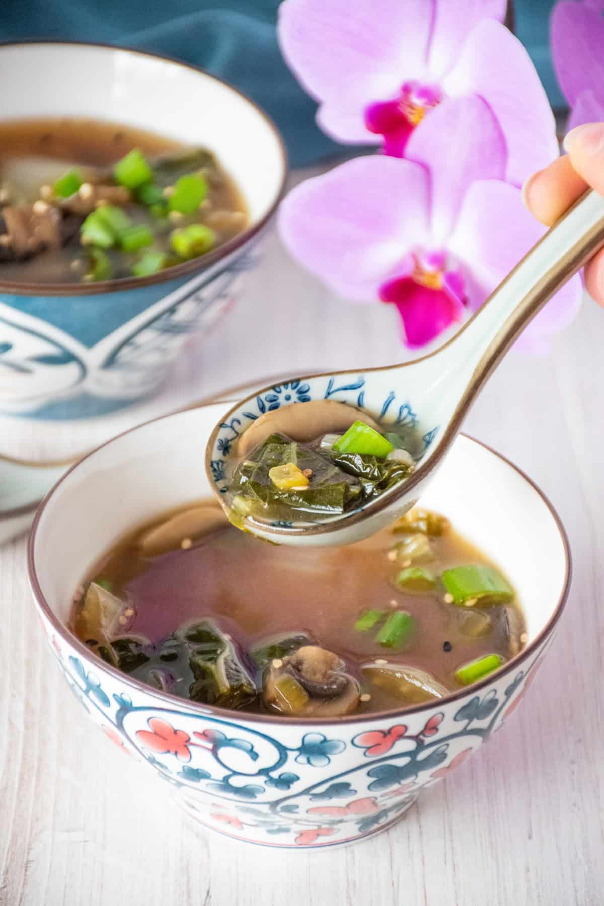 Bowl with bok choy and mushroom soup, garnished with sesame seeds. A spoon is lifting some of the soup forward.