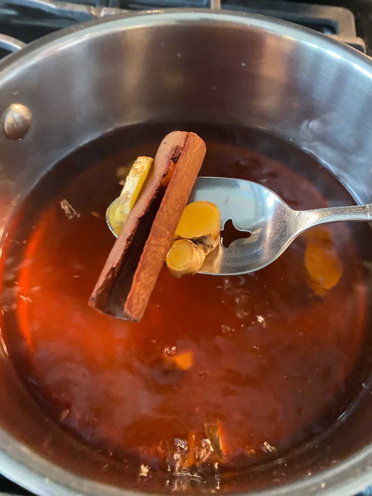 Cinnamon stick and sliced ginger on a slotted spoon, being removed from vegetable broth.