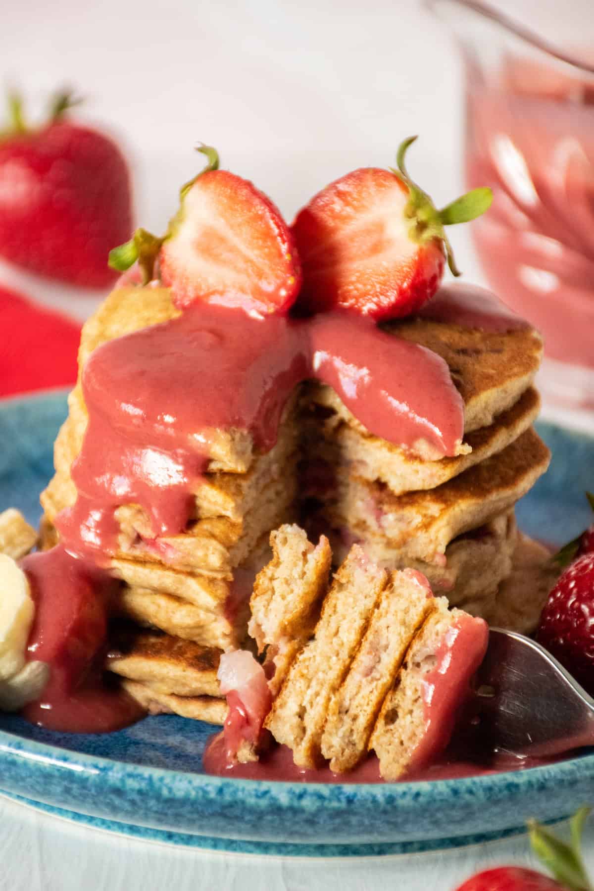 Stack of pancakes with strawberries on top, drizzled with strawberry sauce. A slice is removed to reveal the inside.