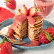 Stack of pancakes with strawberries on top, drizzled with strawberry sauce. A slice is removed from the stack to reveal the inside.