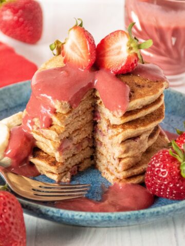 Stack of pancakes with strawberries on top, drizzled with strawberry sauce. A slice is removed from the stack to reveal the inside.