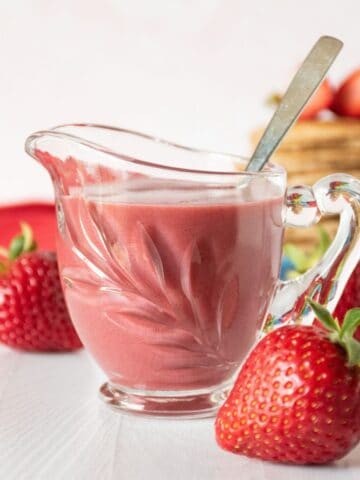Strawberry sauce in a small glass creamer, with fresh strawberries scattered around.